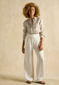 Ľahká, pruhovaná košeľa na gombíky s volánikom, skombinovaná s voľnými bielymi ľanovými nohavicami. Model stojí na textúrovanom povrchu.