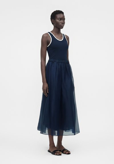 Γυναίκα που φοράει μπλε σκούρο αμάνικο τοπ με λευκές λεπτομέρειες και διάφανο μπλε σκούρο midi φόρεμα με μαύρα σανδάλια, στέκεται μπροστά σε απλό φόντο.