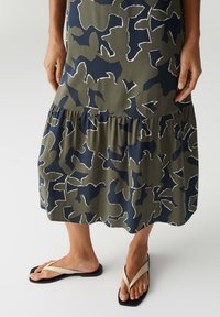 Green and navy patterned tiered dress with white outline, featuring a relaxed fit. Black flip-flops with cream straps and flat soles.