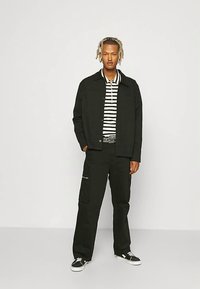 Black jacket over a black and white striped shirt, paired with black cargo pants. Black sneakers. Straight posture against a plain background.