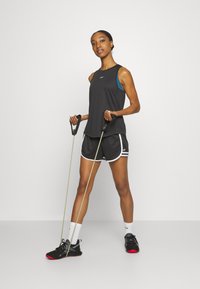Top de treino preto sobre um soutien desportivo azul, combinado com calções pretos com acabamento branco. Segura bandas de resistência e usa sapatos pretos.
