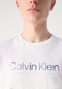 Balts kokvilnas T-krekls ar drosiem ziliem atstarojošiem burtiem, uz kuriem rakstīts "Calvin Klein", un mazāku apakšslogu. Kūniņas apkakles dizains, gluda tekstūra.