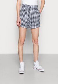 Persona che indossa pantaloni corti a scacchi bianchi e neri con coulisse, una canottiera bianca e sneaker alte bianche, in piedi con le mani nelle tasche.