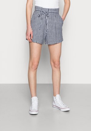 Personne portant un short à carreaux noir et blanc avec cordon de serrage, un débardeur blanc et des baskets montantes blanches, standing avec les mains dans les poches.