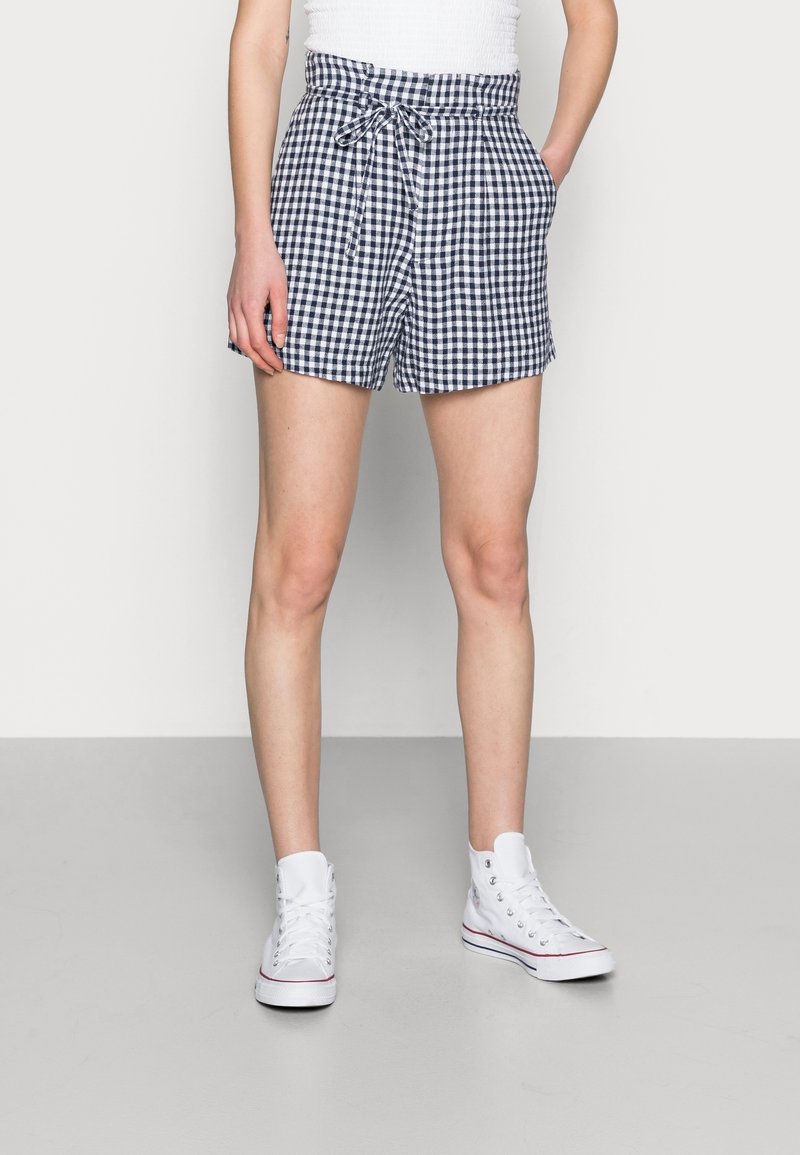 Persona che indossa pantaloni corti a scacchi bianchi e neri con coulisse, una canottiera bianca e sneaker alte bianche, in piedi con le mani nelle tasche.