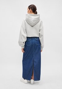 Manga oversized de um hoodie cinza, emparelhado com uma saia longa de jeans azul com dois bolsos traseiros e uma fenda central. Ténis brancos completam o visual.