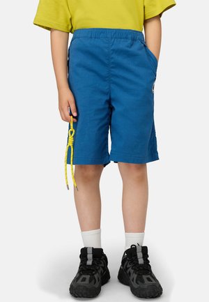 Bambino che indossa pantaloncini blu, maglietta gialla, calzini bianchi e sneakers nere, che tiene in mano un accessorio con una corda gialla.
