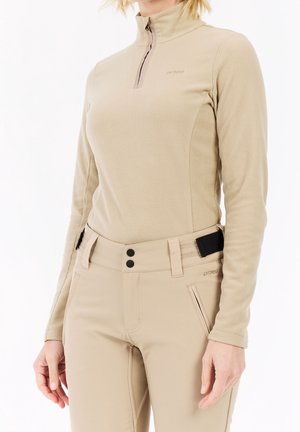 Femme portant un pull en polaire beige et un pantalon beige assorti avec des poches zippées et des bandes latérales noires en velcro.