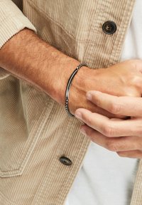 Zwarte en zilveren armband met een glad metalen staaf en een kralenontwerp, gedragen om een pols met een lichtgekleurde corduroy jas.