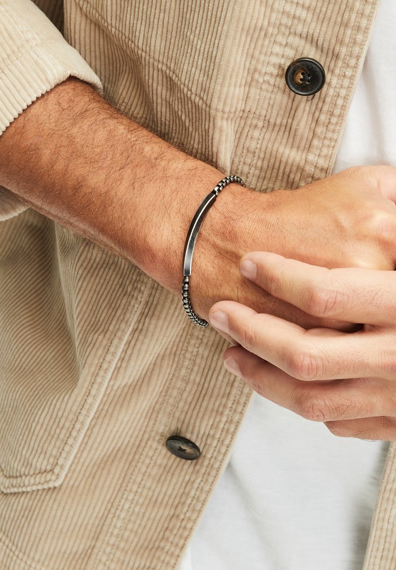 Zwarte en zilveren armband met een glad metalen staaf en een kralenontwerp, gedragen om een pols met een lichtgekleurde corduroy jas.