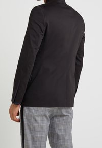 Black tailored blazer with a subtle sheen, featuring a high collar and contrasting inner lining, paired with gray plaid trousers.