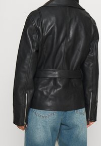 Giacca nera in pelle con vita con cintura, caratterizzata da un ampio colletto e polsini con cerniera. Il retro presenta una trama liscia e una vestibilità su misura.