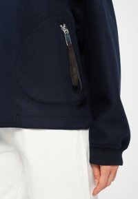 Námořnický fleecový svetr s kapsou na zip a prodlouženými manžetami. Má hladkou texturu a zakřivený šev na kapse.