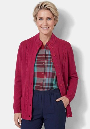 Lächelnde Frau mit kurzen blonden Haaren, die eine rote, geknöpfte Strickjacke über einem karierten Hemd und marineblaue Hosen mit elastischem Bund trägt, steht vor weißem Hintergrund.