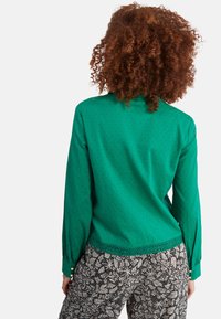 Blouse verte à motif texturé, manches longues et accents de boutons ; associée à un pantalon à motif noir et blanc.