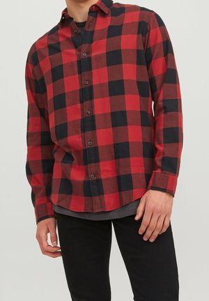 Camisa de franela a cuadros rojos y negros con una tapeta de botones, mangas largas y dobladillo redondeado, llevada sobre una camisa gris.