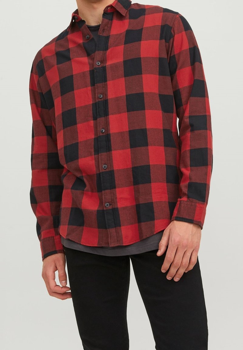 Camicia di flanella a quadri rossi e neri con placca bottonata, maniche lunghe e orlo arrotondato, indossata sopra una maglietta grigia.