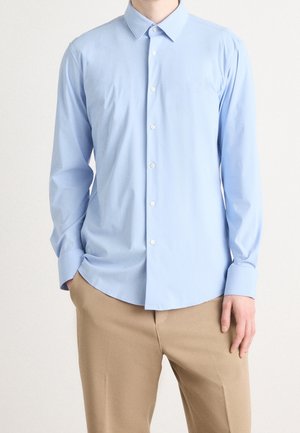 Homme portant une chemise bleu clair à manches longues boutonnée et un pantalon beige, debout avec une main dans la poche sur un fond uni.