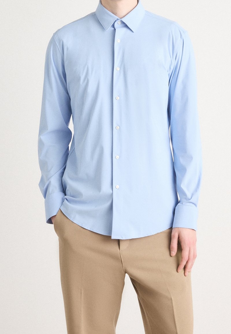 Homme portant une chemise bleu clair à manches longues boutonnée et un pantalon beige, debout avec une main dans la poche sur un fond uni.