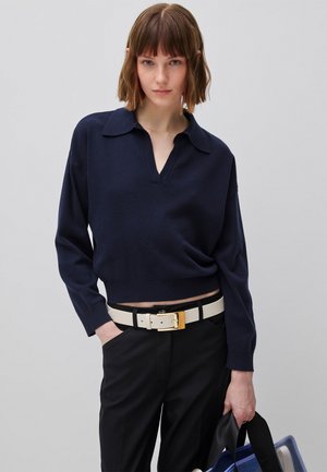 Junge Frau mit kurzen braunen Haaren, die ein marineblaues Langarmshirt, schwarze Hose, weißen Gürtel trägt und eine Tasche mit blau-schwarzen Trägern hält.