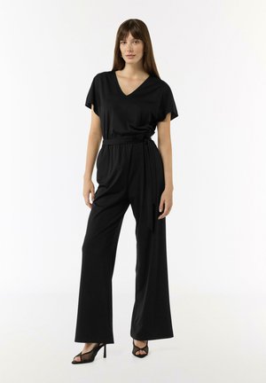 Schwarzer Jumpsuit mit V-Ausschnitt, kurzen Ärmeln und tailliertem Bund. Hat weite Hosenbeine und eine glatte Stoffoberfläche, kombiniert mit schwarzen High Heels.