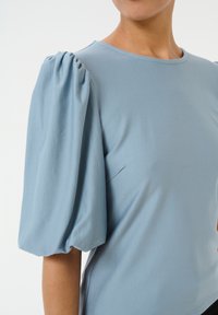 Light blue top featuring a round neckline and voluminous puff sleeves. The fabric appears smooth with subtle texture and structure.