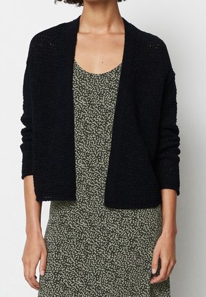 Frau trägt schwarzen, offen gestrickten Cardigan über ärmellosem olivgrünen Kleid mit abstraktem Punktemuster, steht vor einfarbigem Hintergrund.