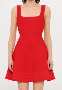 Femme portant une robe rouge sans manches avec un décolleté carré et une jupe évasée, se tenant devant un fond uni.
