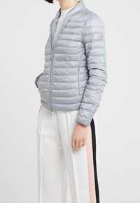 Ljust grå pufferjacka med dragkedja fram, ribbad krage och sidofickor. Har ett horisontellt quiltat mönster och elastiska ärmslut.