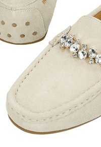 Beige suède instappers met een ronde neus, voorzien van een decoratieve strass-accent en kleine perforaties aan de zijkant.