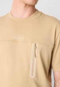 Beige színű, rövid ujjú póló kerek nyakkivágással, elülső cipzáras zsebbel és dombornyomott "HUGO" logóval. Sima, puha szövet textúra.