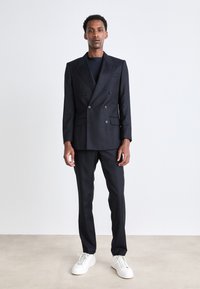 Traje azul marino de doble botonadura con amplias solapas, corte ajustado, pantalones de frente plano y zapatillas blancas. Textura suave, diseño minimalista.