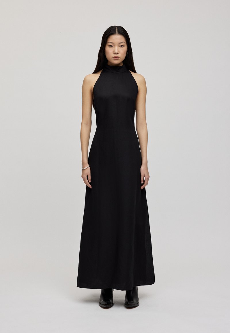 Rochie maxi cu gât halter de culoare neagră, realizată dintr-un material neted, cu un corset ajustat și o fustă dreaptă, fluidă. Pur tată cu cizme negre până la gleznă.