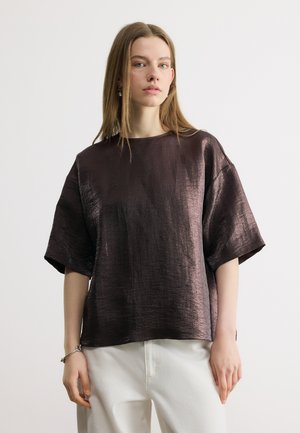 Junge Frau mit glattem, hellbraunem Haar, die eine lockere, kurzärmelige dunkle metallische Bluse und weiße Hose trägt und vor einem schlichten hellen Hintergrund steht.