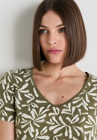 Camiseta de algodón verde oliva con cuello en V y mangas cortas, que presenta patrones de impresión botánica blanca y un acabado de tela texturizada.