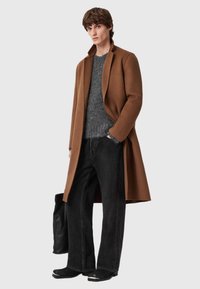 Smeđi vuneni kaput preko sivo pletenog džempera, uparen s crnim širokim trapericama i crnim čizmama do članka s srebrnim detaljima. Jednostavna pozadina.