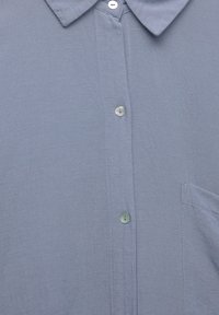 Chemise bleu clair à boutons avec un col, présentant une petite poche et un tissu lisse et texturé. Quatre boutons visibles.