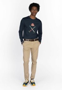 Junger Mann stehend mit Händen in den Taschen, trägt ein navyblaues Langarmshirt mit einem Grafik von gekreuzten Schlägern und einem Helm, khakifarbene Hose und mehrfarbige Sneakers.