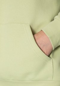 Ljust grönt sweatshirt med mjuk textur, med en framficka och ribbade ärmslut. Materialet verkar elastiskt och bekvämt.