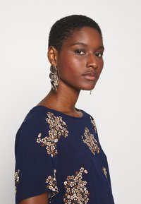 T-shirt azul marinho com mangas curtas, adornado com detalhes em lantejoulas florais em padrões brancos, dourados e bronze. Grandes brincos de prata.
