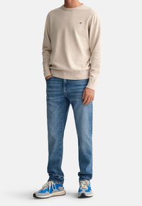 Pull beige en maille, jeans bleus et baskets multicolores. Le pull présente un col rond et une broderie de logo discrète ; les jeans ont une coupe classique.