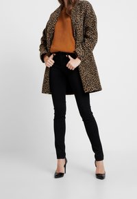 Abrigo de estampado de leopardo, suéter naranja de canalé, jeans ajustados negros y tacones negros. El abrigo tiene un cuello y bolsillos laterales. Texturas suaves.