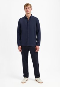 Man in een donkerblauwe lange mouwen overhemd met knopen, bijpassende donkerblauwe broek en witte sneakers, staande tegen een witte achtergrond.