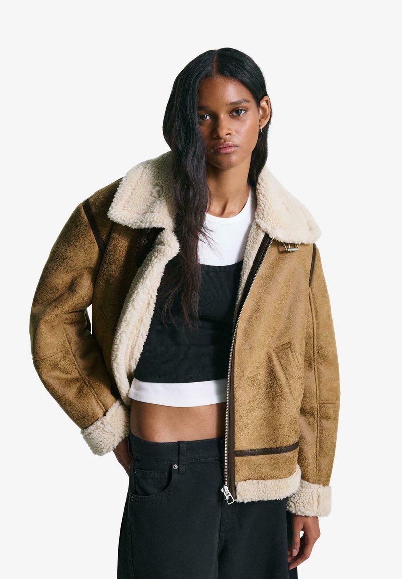 Brun shearling bomberjacka med en krämfärgad krage och manschetter, med blixtlås och mörka accenter. Bärs över en svart topp och jeans.