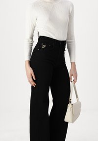 Pantaloni neri a vita alta con un design aderente, dotati di tasca laterale e dettagli in metallo argentato. Abbinati a un dolcevita bianco a coste e a una borsa leggera.