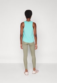 Jasnoniebieski bezrękawnik z zaokrąglonym dołem, zestawiony z oliwkowymi legginsami. Modelka w jasnoróżowych trampkach. Minimalistyczne tło.