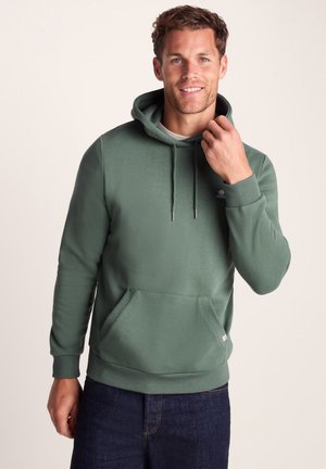 Man wearing green hooded sweatshirt with front pocket and drawstrings, standing against plain light background.