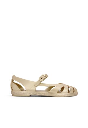 Sandalia plana beige con puntera abierta, laterales recortados y correa con hebilla, mostrada de perfil sobre un fondo blanco liso.