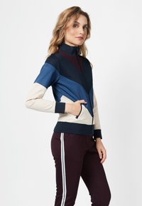 Kleurgebloekte ritsjack met marineblauwe, blauwe, bordeauxrode en crèmekleurige secties. Beschikt over een hoge kraag en zijzakken. Gladde textuur.