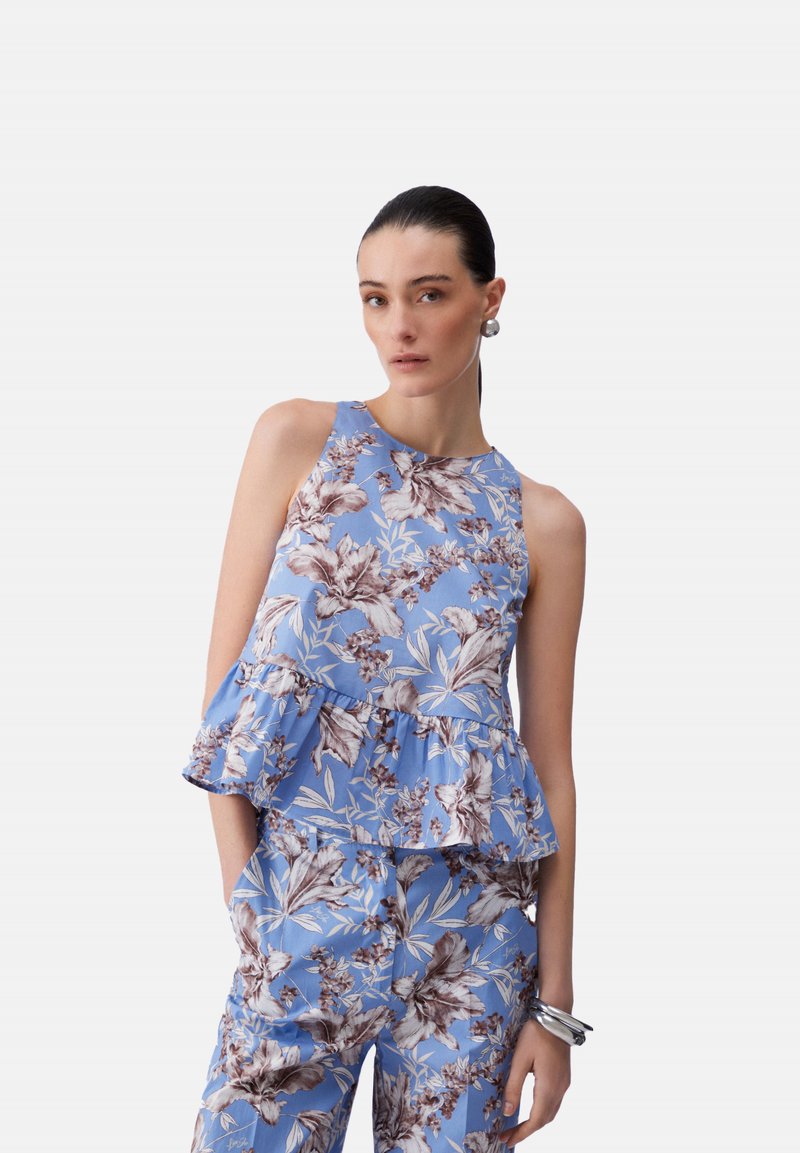 Femme aux cheveux tirés en arrière portant un haut sans manches bleu à motif floral et un pantalon assorti, accessoirisée avec des boucles d'oreilles et des bracelets argentés.